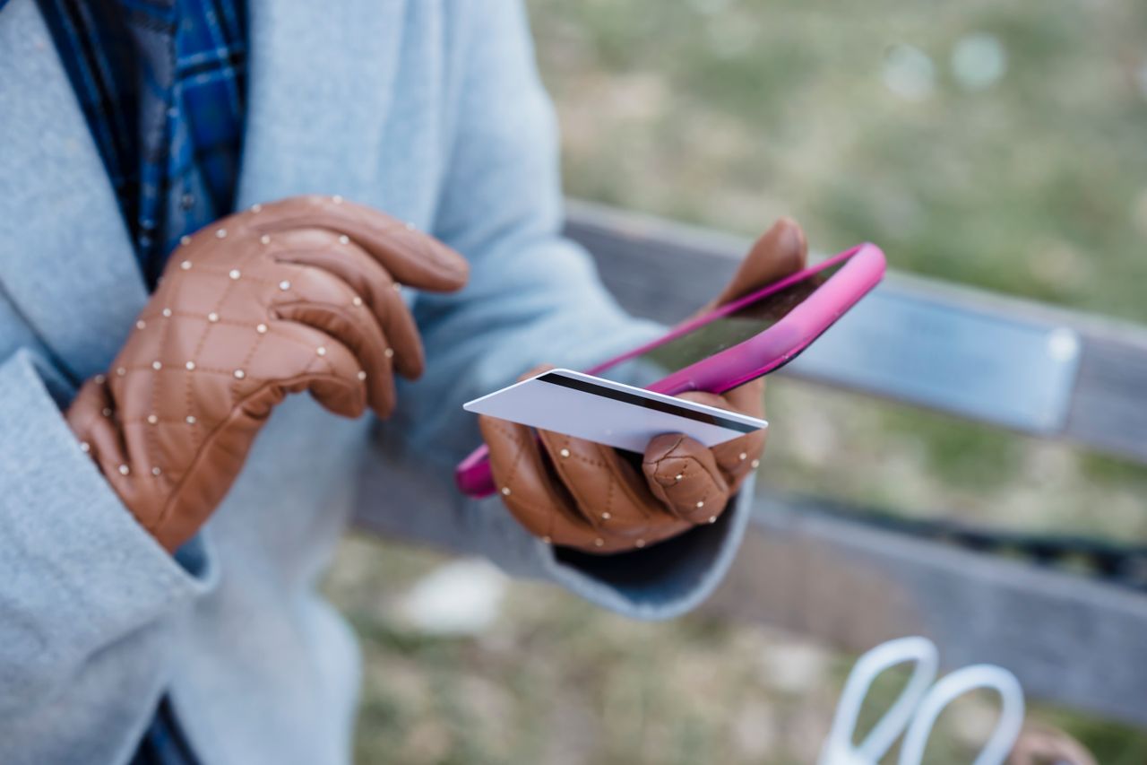 Person in gloves holding a bank card and a smartphone while paying for something online on a park bench