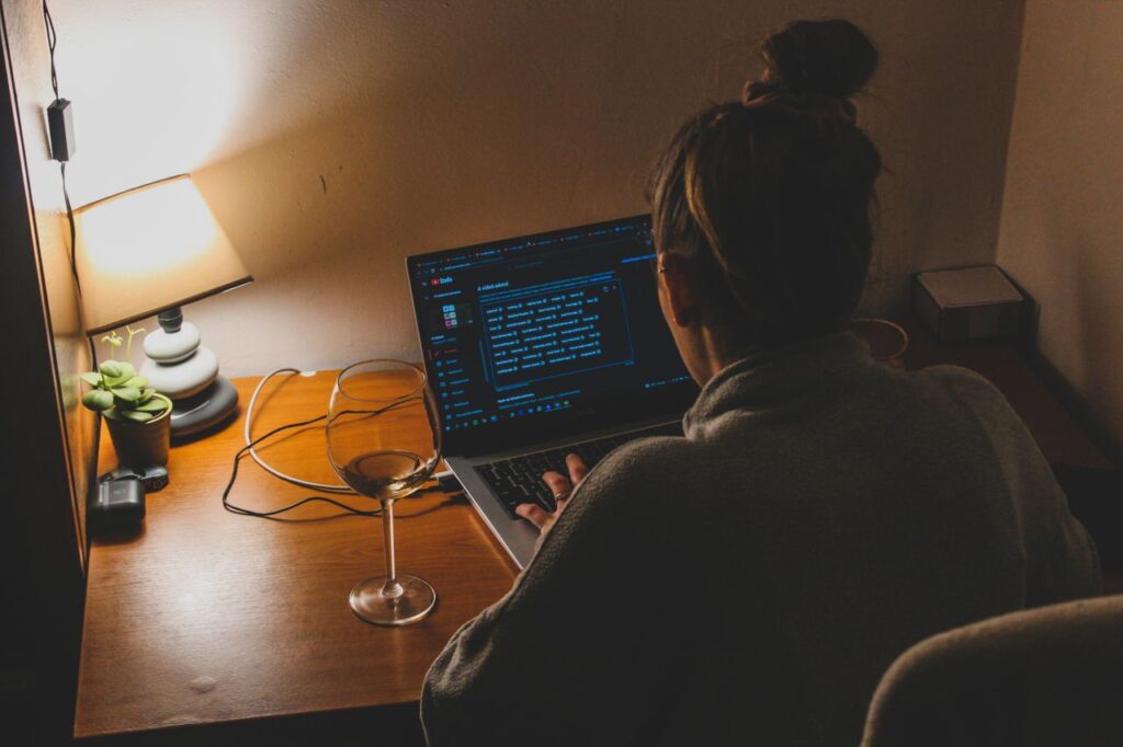 Person seen from behind working late at night on a laptop at a small desk lit by a lamp