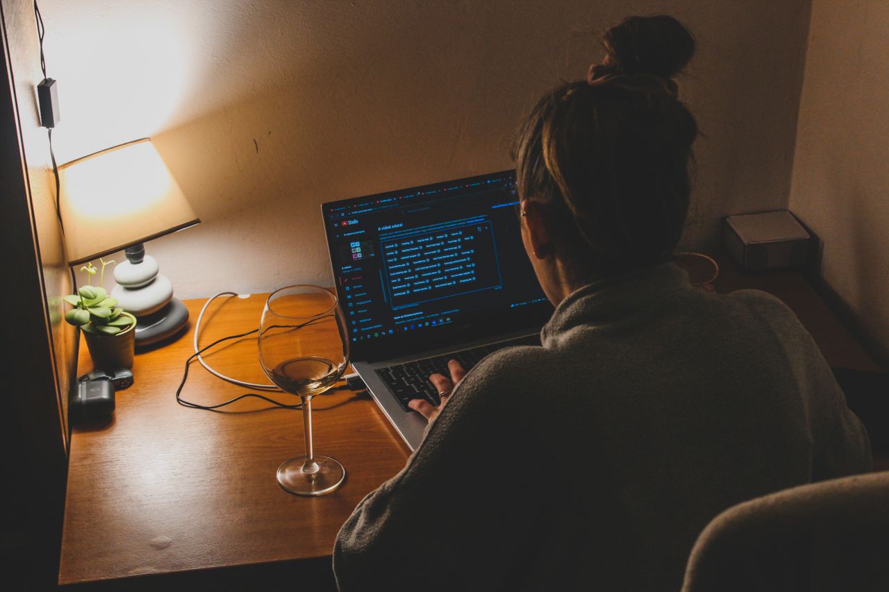 Person seen from behind working late at night on a laptop at a small desk lit by a lamp