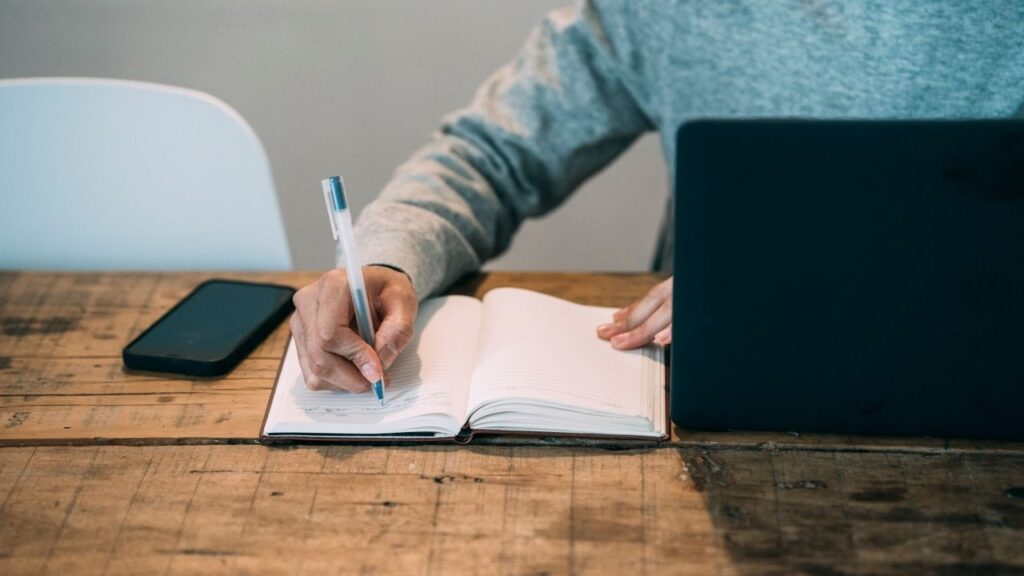 Man writing in a notebook at a wooden table beside a laptop and a smartphone