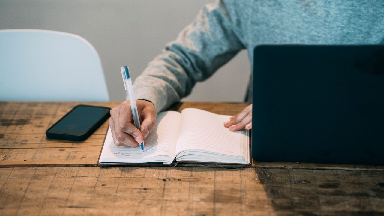 Home 4 Man writing in a notebook at a wooden table beside a laptop and a smartphone